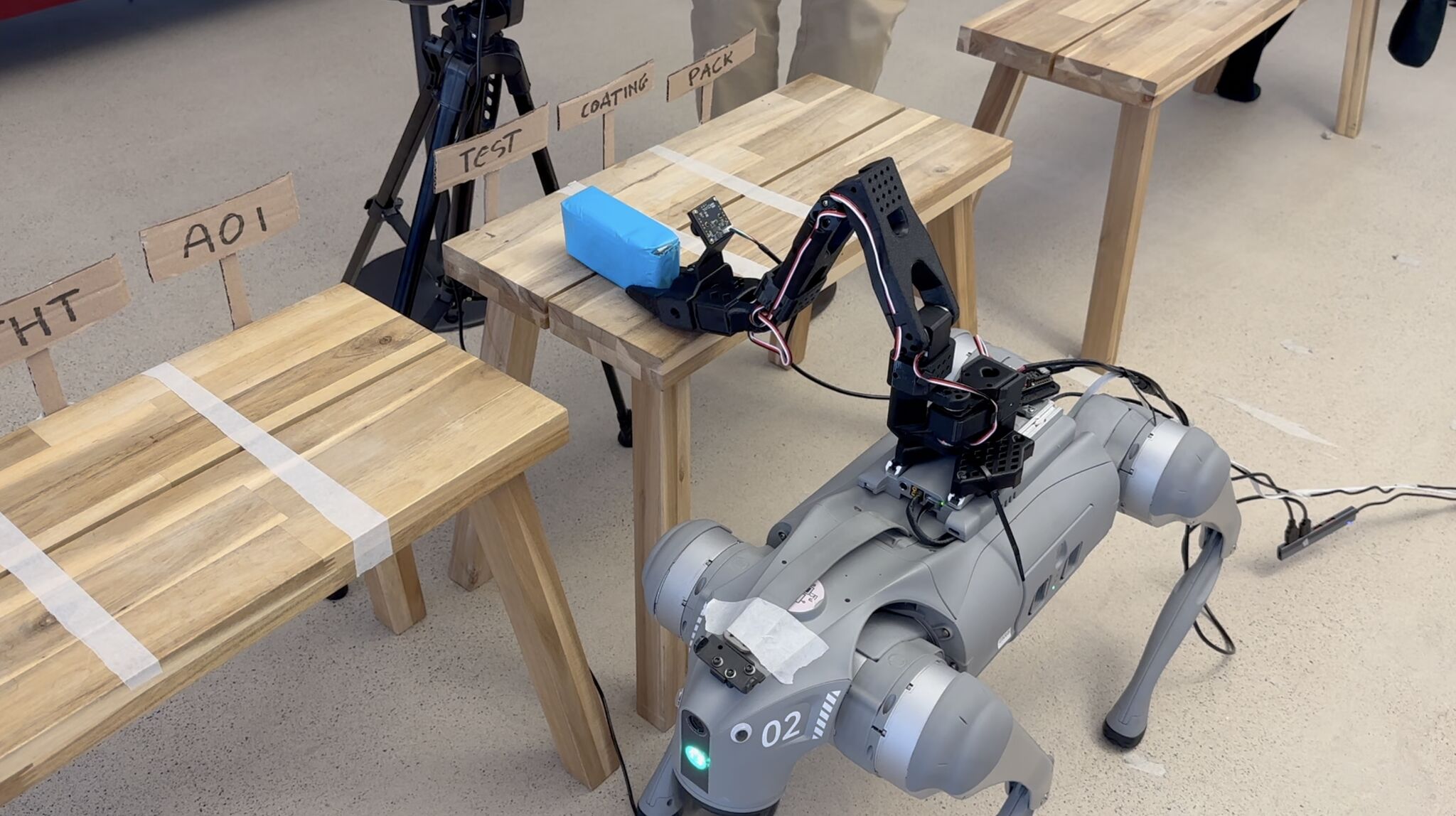 A robot dog with a mounted arm picking up a part from a mock assembly line of wooden benches labelled AOI, TEST, COATING and PACK.